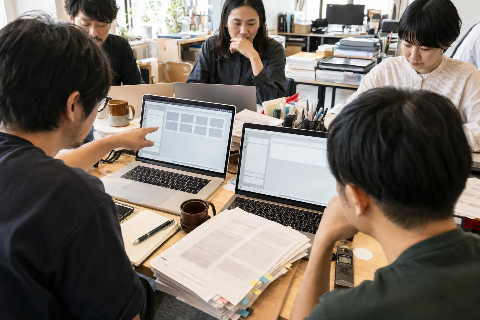 小さな職場のテーブルで、複数人がノートパソコンを使い、実際の書類や議事録をAIで処理している様子の写真調イラスト。